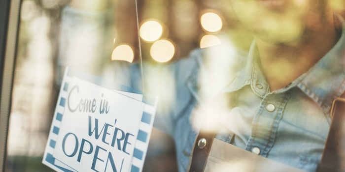  Person in denim jacker hanging a "Come in WE'RE OPEN" sign on a glass door
