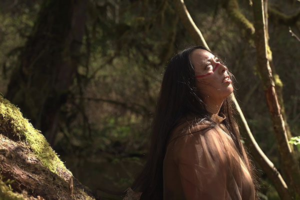 A Mexican woman with long black hair and a strip of red facepaint under her eyes stands in the forest. She's looking upward and natural light bathes her face.