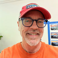 A white man wearing a baseball cap and glasses, with a short white beard