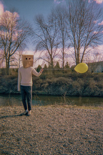 A figure wearing a sweater and a paper bag over their head holds a yellow balloon by a string. It's impossibly stretched taut to the side.