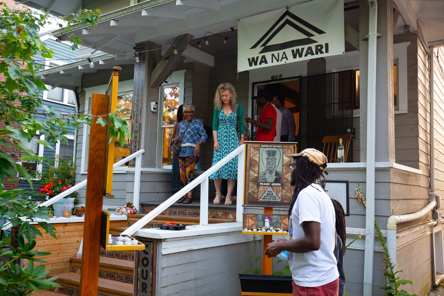 A house with a banner above the doorway that says "Wa Na Wari". People are exiting the house. People are seen in the foreground walking towards the house.