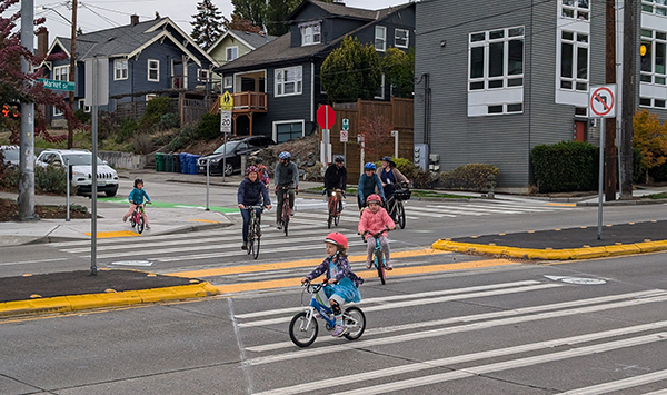 Kids and adults biking through a very large and well marked crosswalk with 