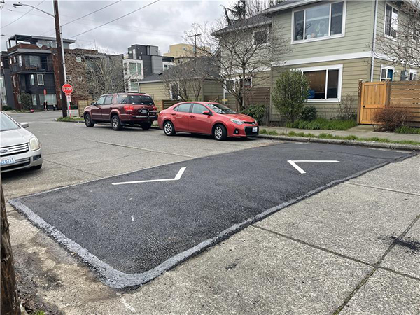 Picture of a street with a dark black speed hump across it with parked cars and houses in the background