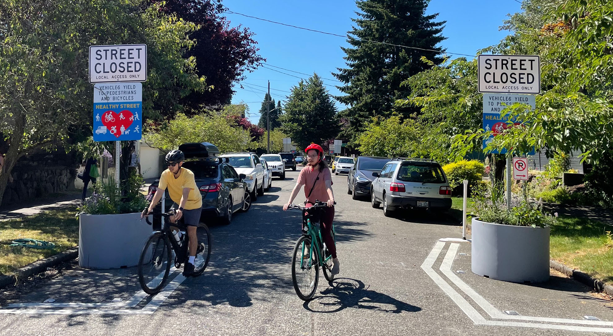 Two people on bikes on a Healthy Street with signs in planters