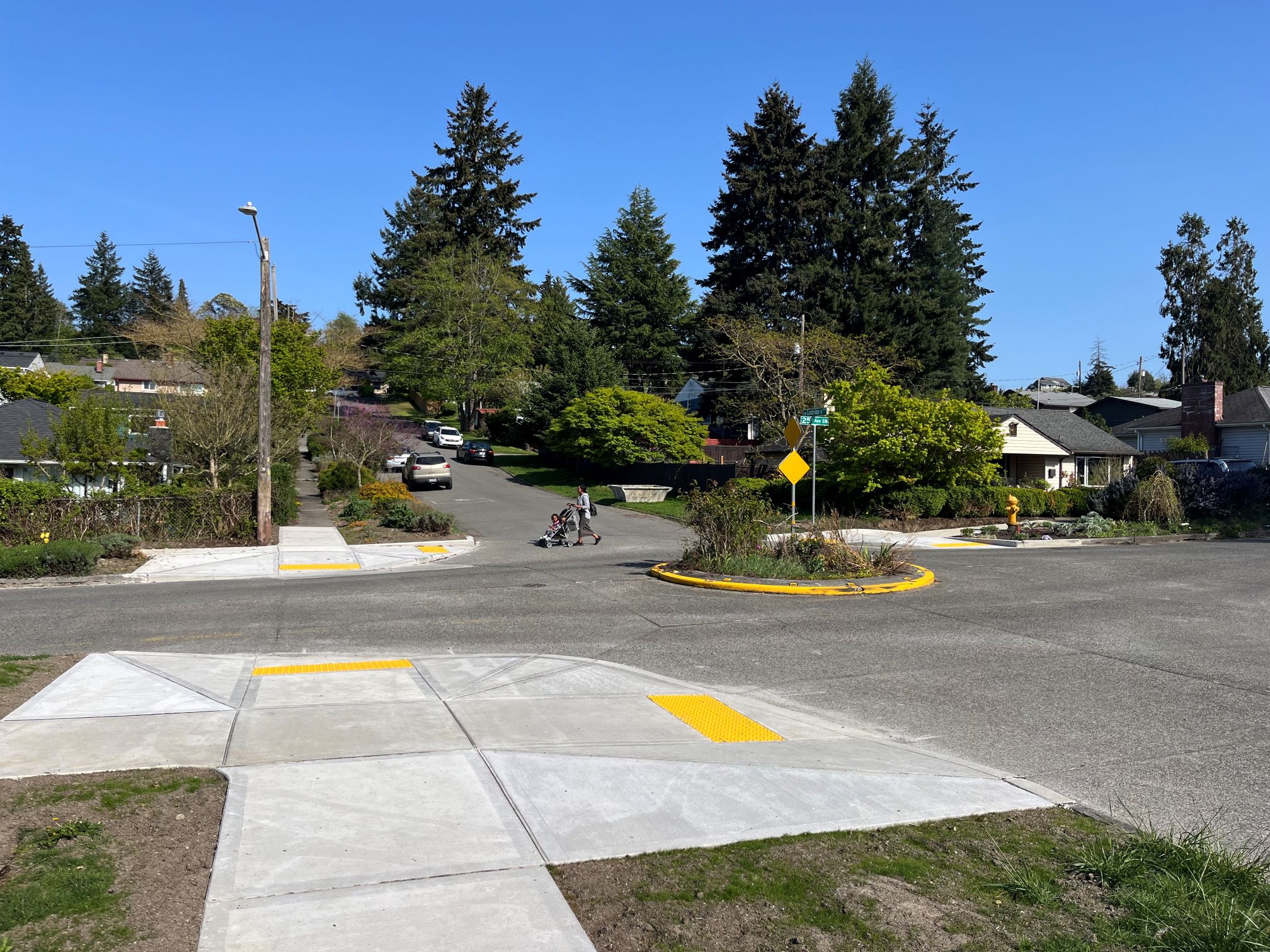 Intersection of 25th Ave SW and SW Cambridge St showing new curb ramps with tactile yellow strips, a roundabout with greenery, and a person pushing a stroller across the street under a clear blue sky.