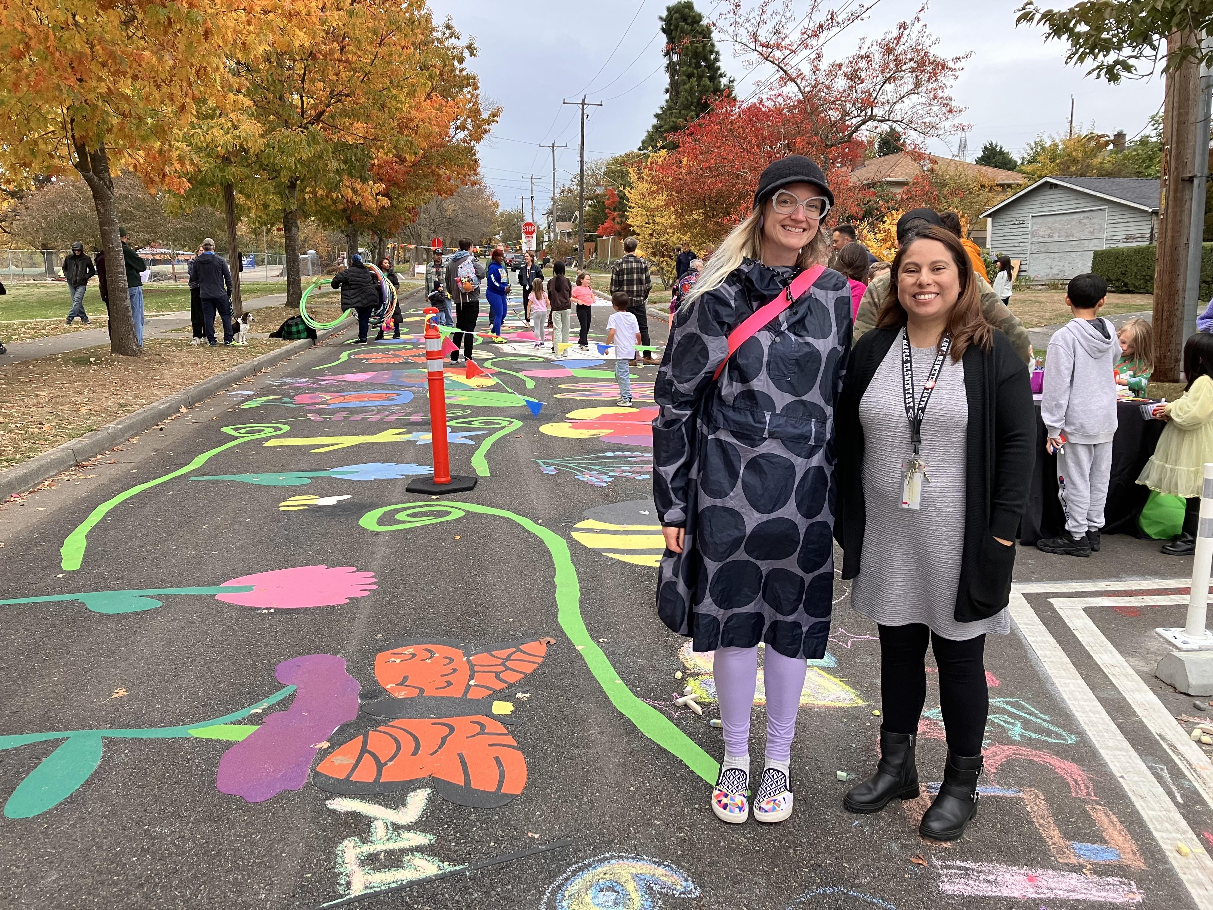 Two adults stand on a road with colorful artwork on it, while kids play in the background.