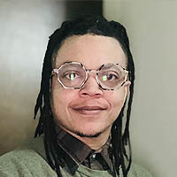 Dorian has black hair in short dreadlock, is wearing glasses and has a green sweater