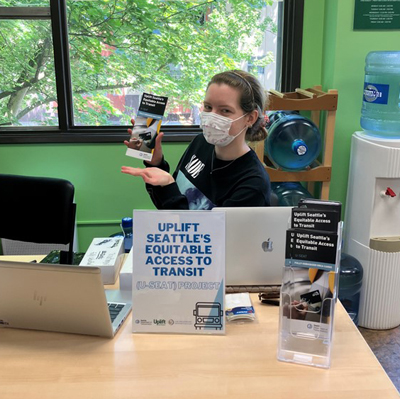 A person sitting behind a desk with a sign that says "Uplift Seattle's Equitable Access to Transit" and they are holding up a brochure. 