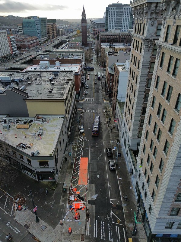 A drone shot overhead, facing southbound down 3rd Ave from Yesler Way. An orange metal plate and safety cones surround an area where repaving is happening just south of Yesler