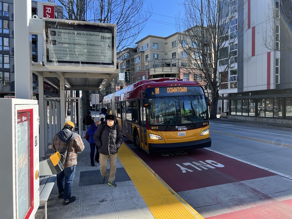 Passengers wait at a Madison St RapidRide station as a King County Metro G Line bus approaches, displaying "Downtown" as its destination.