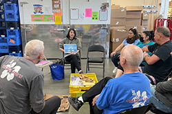 A person addresses several people seated in a grocery warehouse.