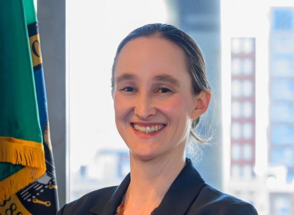 Mayor Wilson smiling, in a dark blazer stands with her arms crossed in an office setting, with two flags behind her and a city skyline visible through the windows