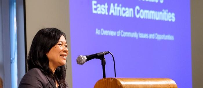 Close up of woman speaking at a podium with a blurry screen behind her saying East African Community.