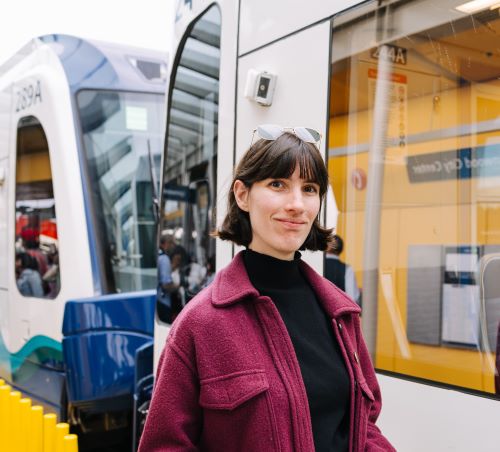 Portrait of Rebecca smiling slightly at the camera in a magenta coat with a light rail train in the background.