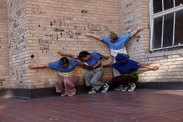 Four dancers lean against a brick wall with graffiti, arms extended and heads down.