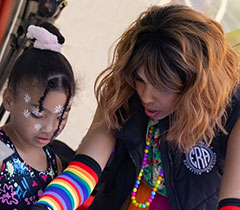 Kay wears long gloves with rainbow stripes and helps a young Black girl with an art project