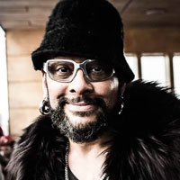 Ari wears a tall bucket hat, chunky shades, and a black coat with a heavy, furry collar.