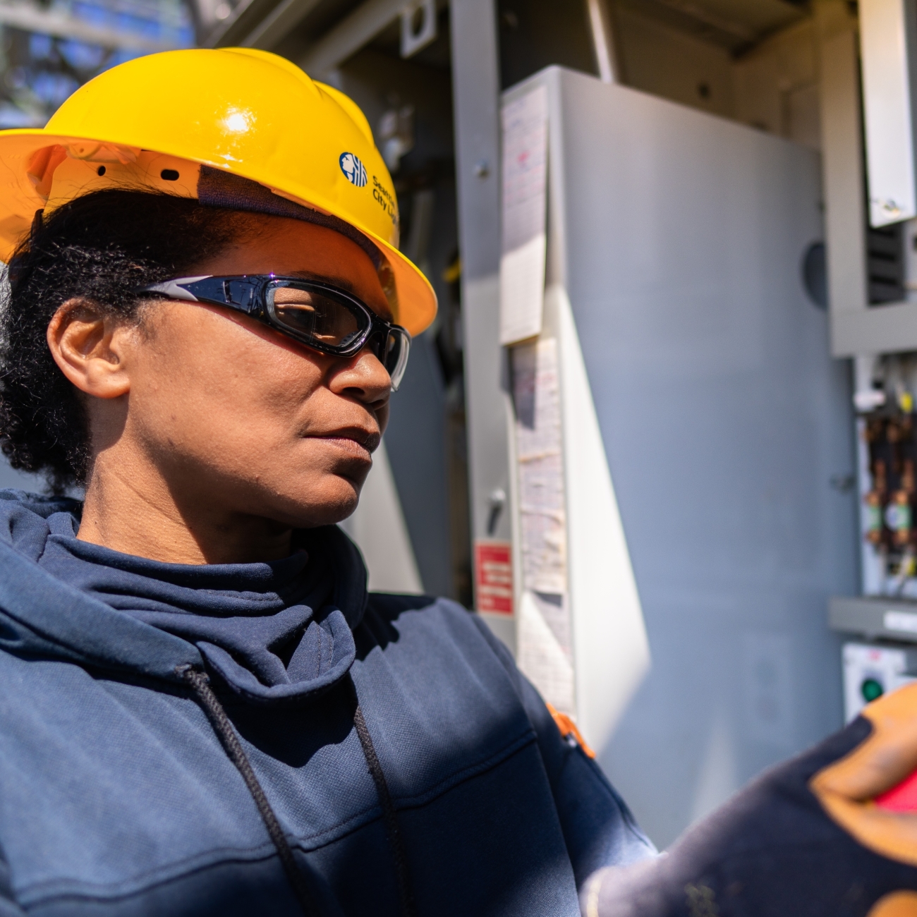 A woman wearing a yellow hard hat and a dark blue work jacket while examining an open electrical panel. The individual is an electrician engaged in maintenance or inspection work. The background is blurred, emphasizing the focus on the person and the task at hand.
