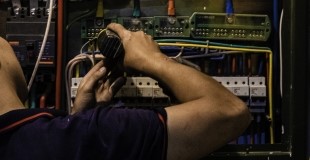 A person is working on an electrical panel, connecting wires. The individual’s focus is on the task at hand, with their hands manipulating cables within the open panel which contains various electrical components.