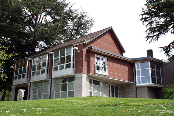 A red brick 2 story building with many windows, situated on a hill with trees around it.