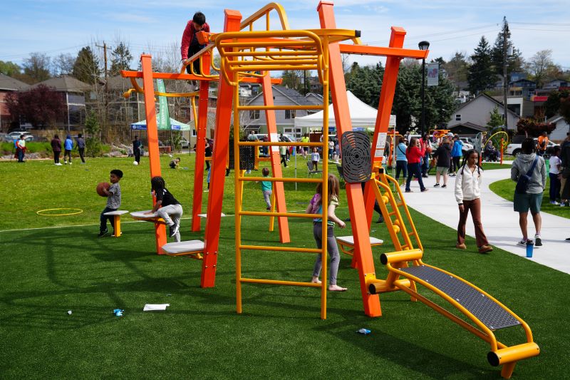 Children play and climb on a vibrant orange and yellow fitness structure in a grassy area of the park. Families and children are scattered throughout, with tents and more play areas visible in the background.