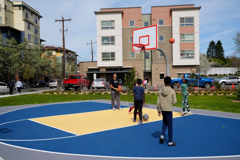 Children and a young adult shoot basketballs on a newly painted half basketball court at Cheryl Chow Park. Apartment buildings and parked cars line the background, and the scene is lively with activity and greenery.
