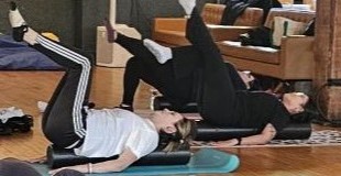 Two participants do a Tabletop position on a lengthwise foam roller during an indoor Pilates class