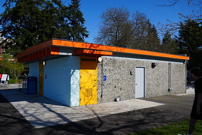 A small flat-roofed building with orange trim, yellow doors, and textured gray walls sits along a paved path