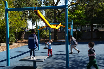 Several children run and play on blue and orange playground zipline structure on a blue rubber surface.