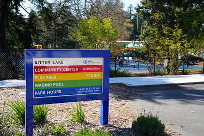 A colorful outdoor sign reads “Bitter Lake Community Center” with listings for play area, wading pool, tennis, and park hours, with the playground visible in the background.