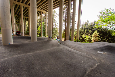 A child rides a scooter across the smooth asphalt surface of the I-5 Colonnade pump track beneath the freeway overpass.