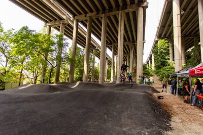 Cyclists ride a paved pump track under the I-5 Highway, with tall freeway columns and event tents in the background.