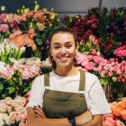 Florist in apron smiling and standing in front of flowers with arms crossed.