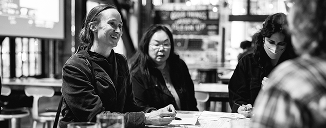 Mayor Katie B. Wilson along with three people sit at a table in a café or public space, smiling and talking while writing on papers, with other people and signage visible in the background.